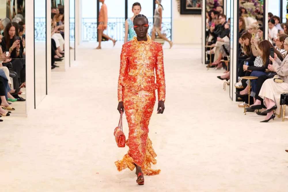 Model wearing vibrant orange and red patterned long-sleeve dress walking down white runway at Chanel Cruise 2026/27 show with seated audience on sides